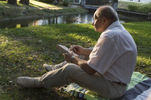 An elderly man in a white shirt is sitting on a blanket, on the ground in a park and reading an interesting book. A pensioner alone is resting in nature, passionate about his hobby